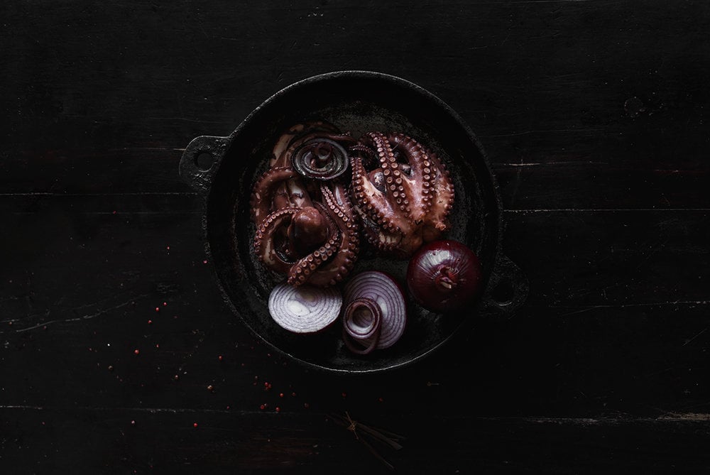 Coqui Coqui Papholchac, Mexico — Overhead view of octopus tentacles and sliced red onions in dark cast-iron pan, moody culinary still life.