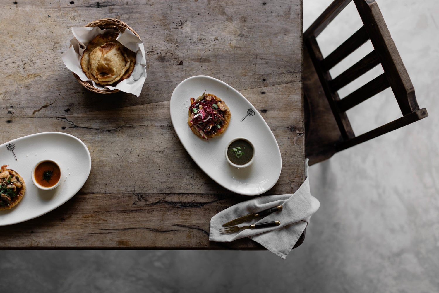 Coqui Coqui Papholchac, Mexico — Overhead view of plated appetizers with beetroot and garnish, bread basket, and cutlery on weathered wood table with sculptural black chair.