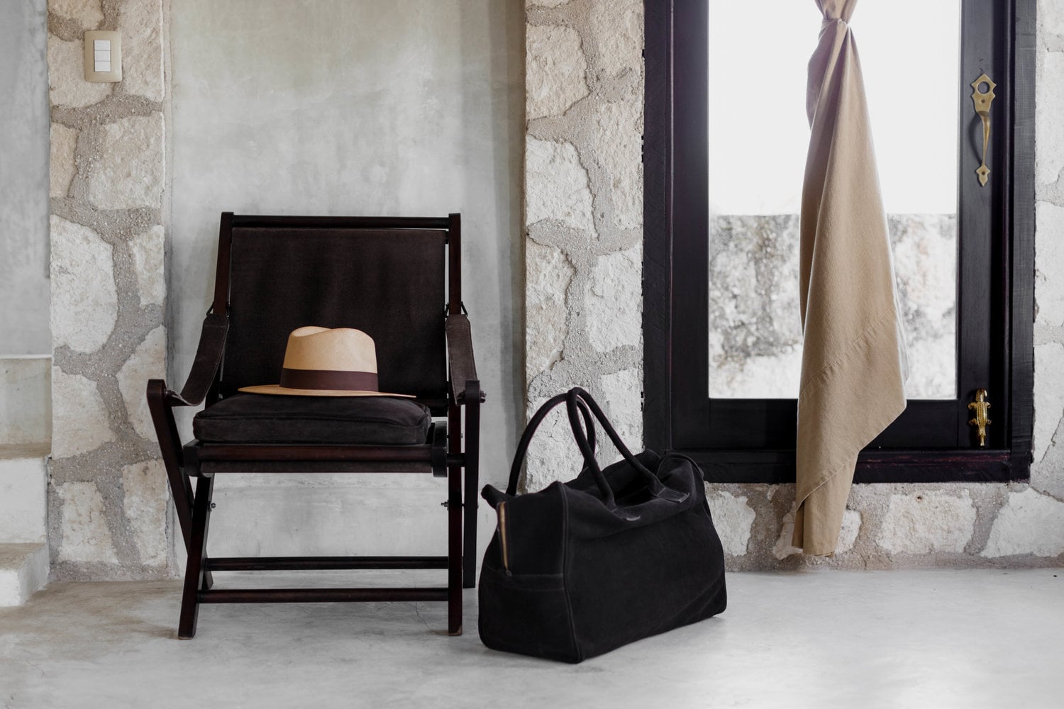 Coqui Coqui Papholchac, Mexico — Black wooden chair with straw hat and leather bag beside cream linen curtain, stone walls, minimalist coastal interior.