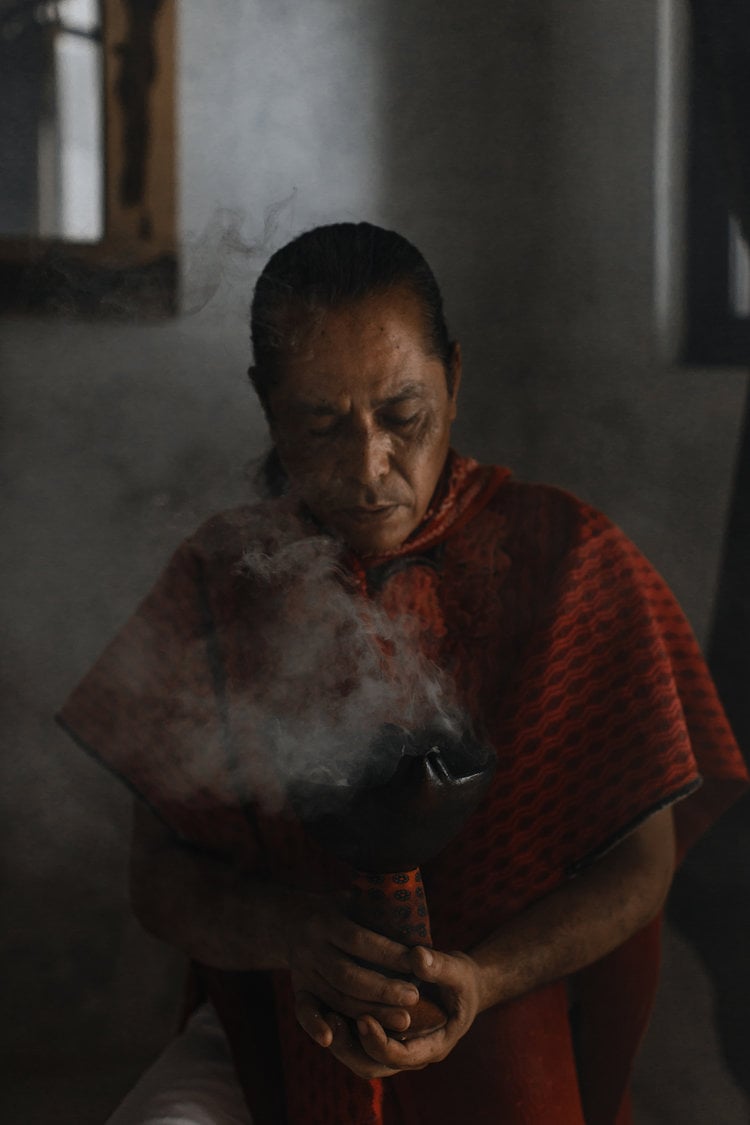 Coqui Coqui Papholchac, Mexico — Woman in red striped shirt holding smoking ceramic vessel in minimalist whitewashed interior with natural light.
