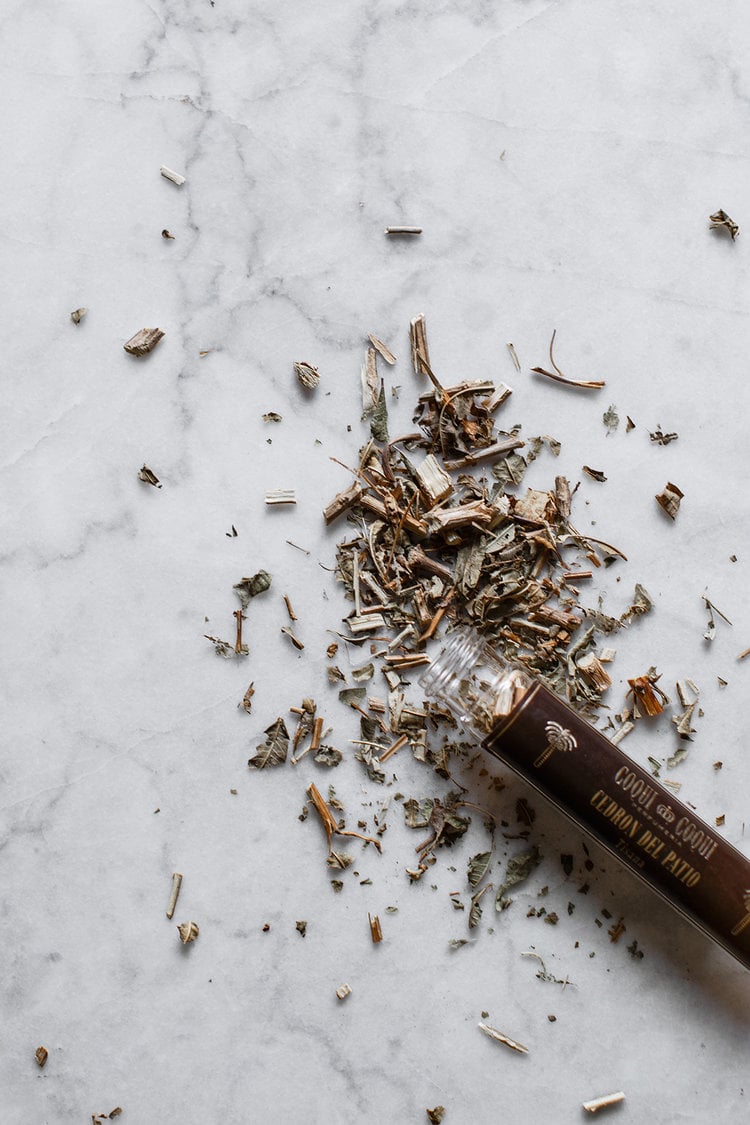 Coqui Coqui Papholchac, Mexico — Dried herbs and botanicals spilling from dark glass bottle onto white marble surface, artisanal wellness aesthetic.
