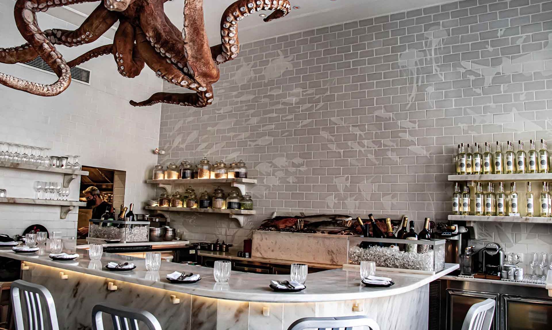 A Cevicheria, Portugal — Bronze octopus sculpture above marble counter with subway tile walls, candles, and olive oil bottles in luxury seafood restaurant.