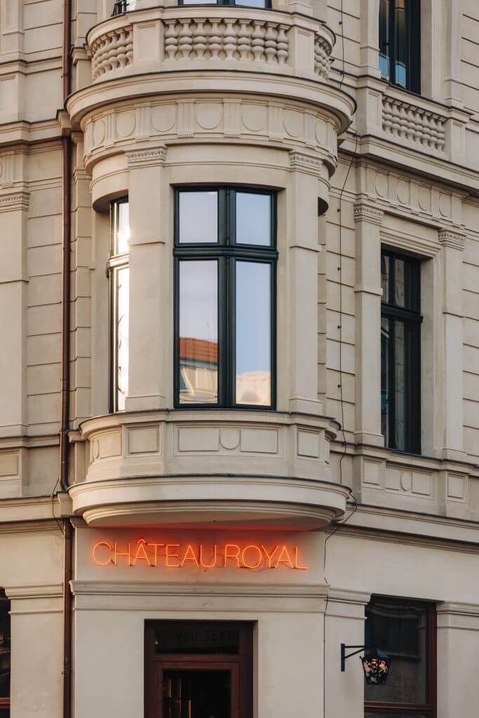 Château Royal, Europe — Cream stone corner tower with ornate balustrade, dark-framed windows, and glowing neon signage on historic Haussmann-style facade.