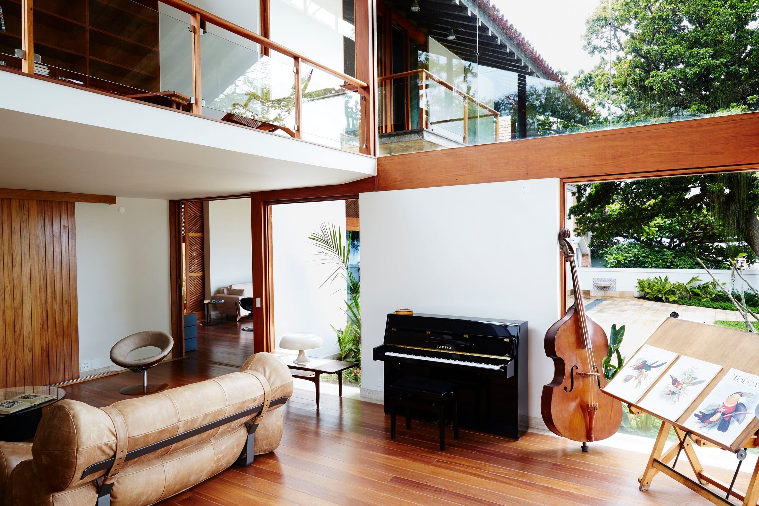 Chez Georges, South America — Double-height living room with black piano, cello, leather sofa, wooden flooring, and floor-to-ceiling glass overlooking lush gardens.