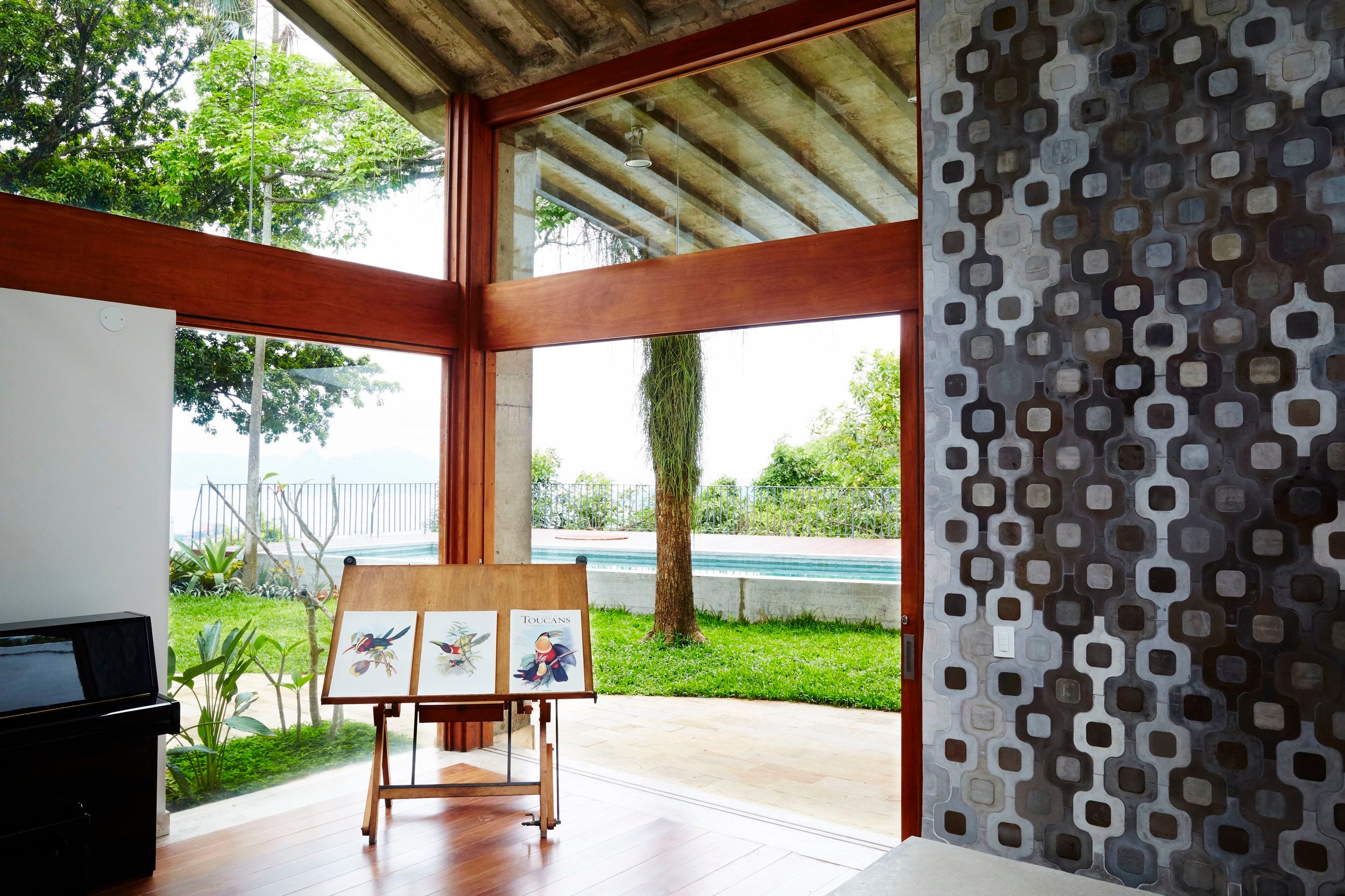 Chez Georges, South America — Modernist gallery space with wood-framed glass walls, concrete ceiling, patterned stone accent wall, and pool views through lush landscaping.