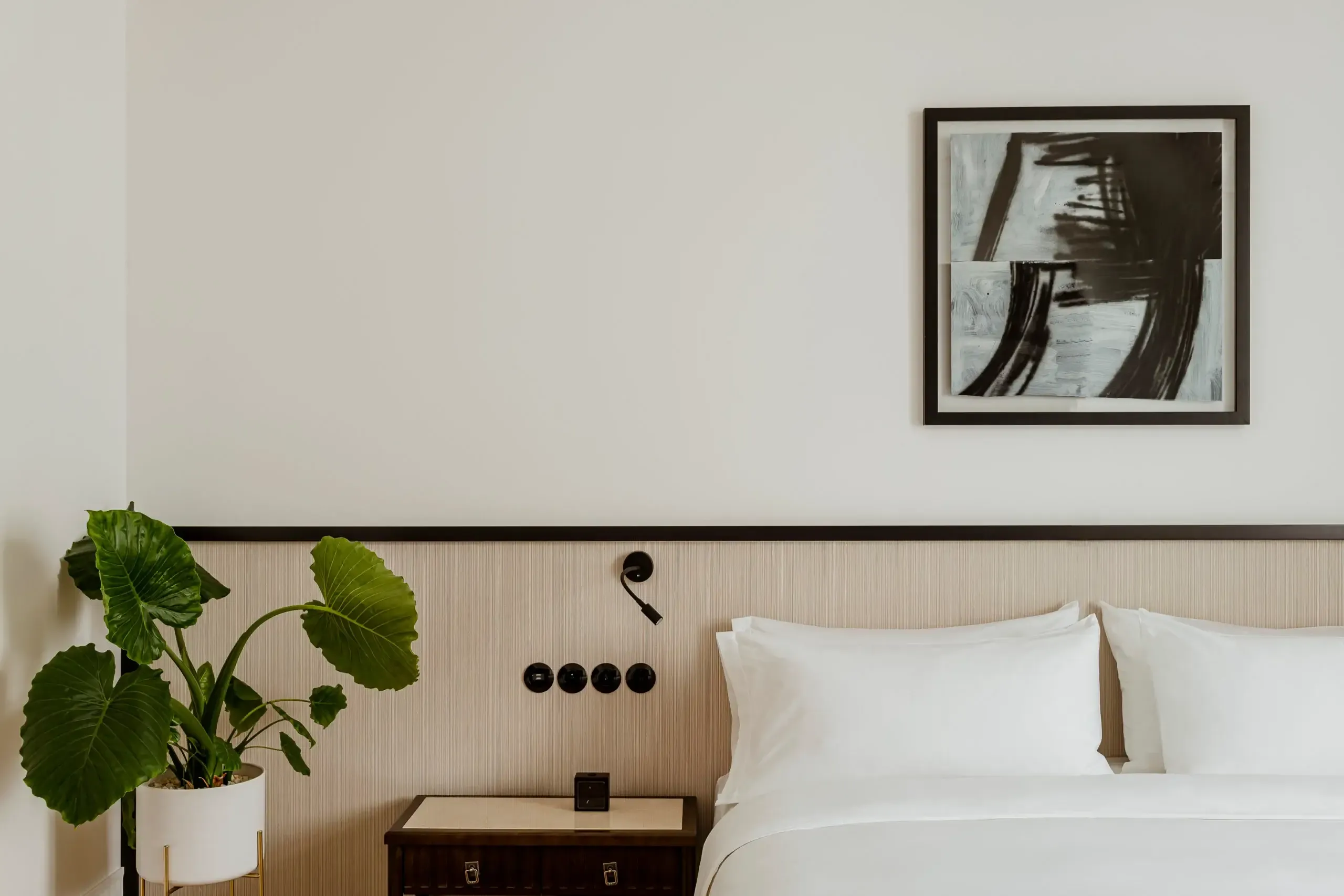 Nobu Warsaw, Poland — Modern hotel bedroom with minimalist black headboard, white linens, potted plant, abstract framed artwork, and warm neutral palette.