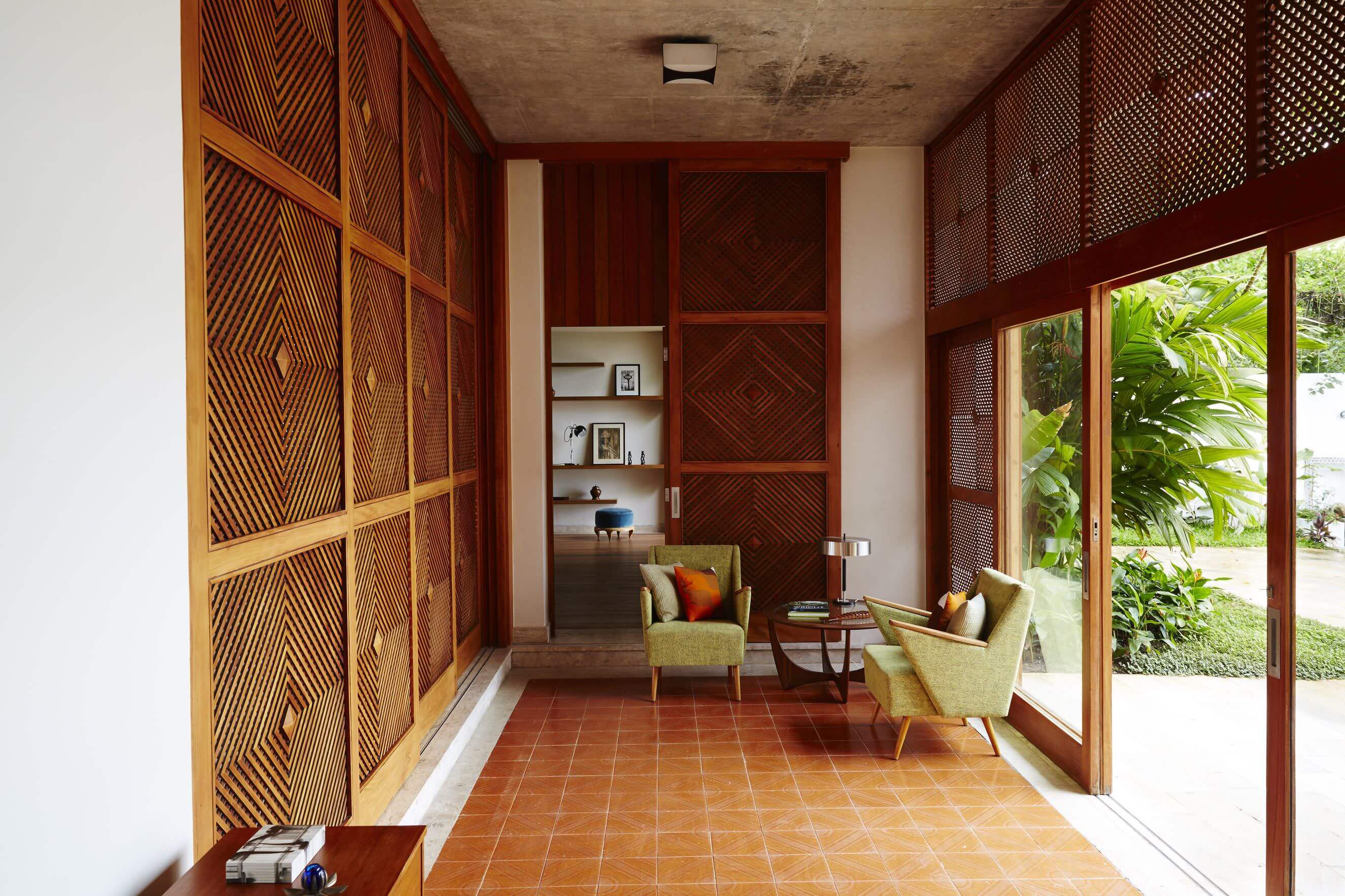 Chez Georges, South America — Covered veranda with geometric wooden screens, terracotta tile flooring, chartreuse mid-century chairs, and garden views through glass doors.