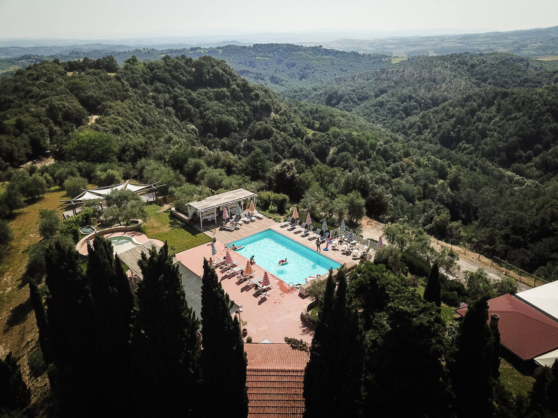 Villa Lena, Tuscany, Italy — Clifftop swimming pool overlooking forested valleys, with cypress trees, loungers, and terraced architecture nestled in rolling hills.