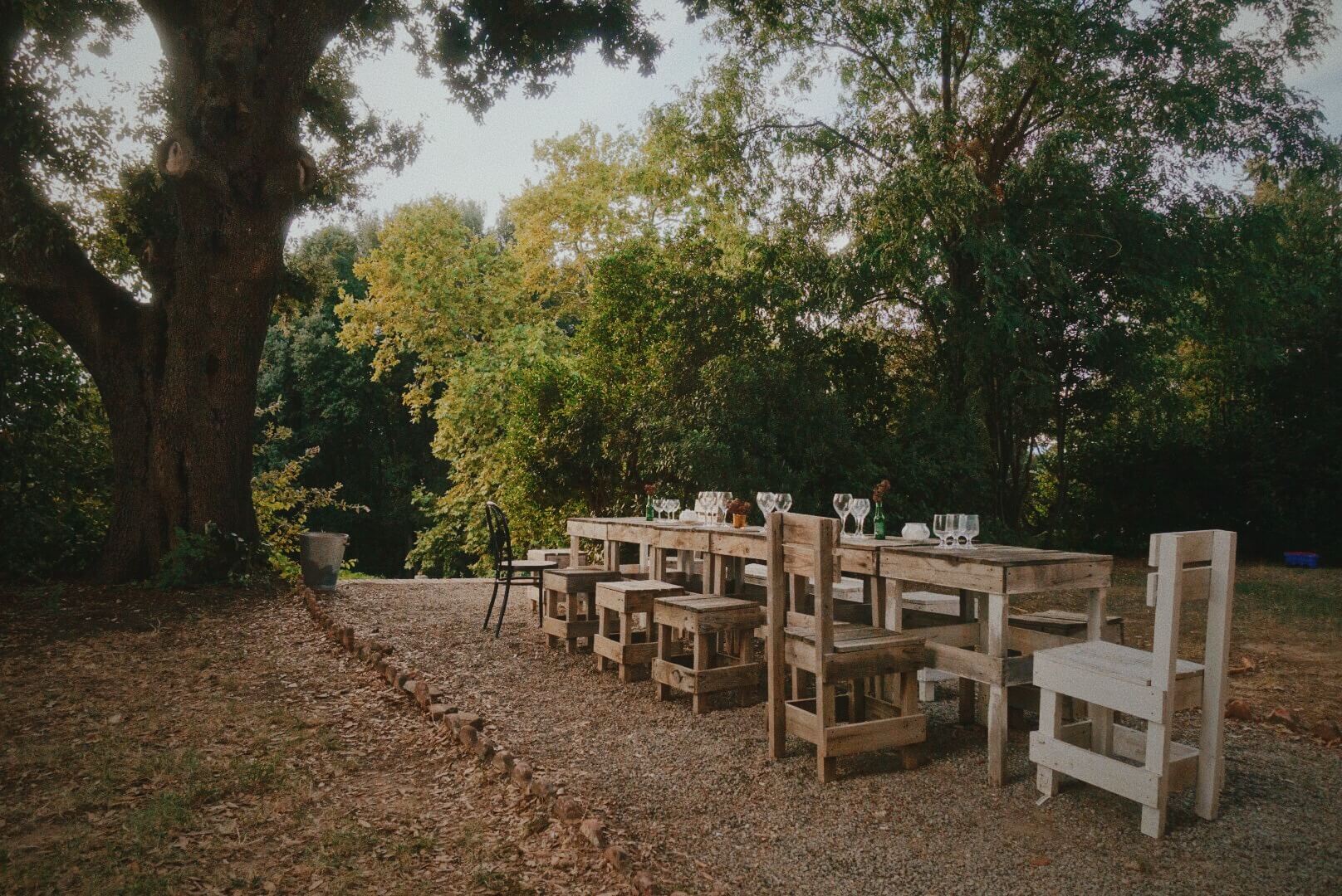 Villa Lena, Europe — Wooden dining tables and chairs arranged on gravel beneath mature trees in autumn, set for outdoor entertaining with glassware and table settings.