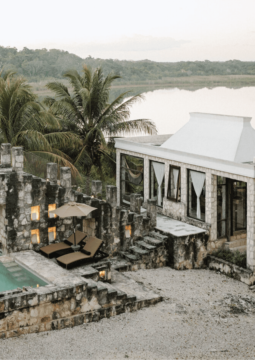 Coqui Coqui Papholchac, Mexico — Stone pavilion with white roof overlooking lagoon and jungle, palm trees, lounge chairs, candlelit terrace at dusk.