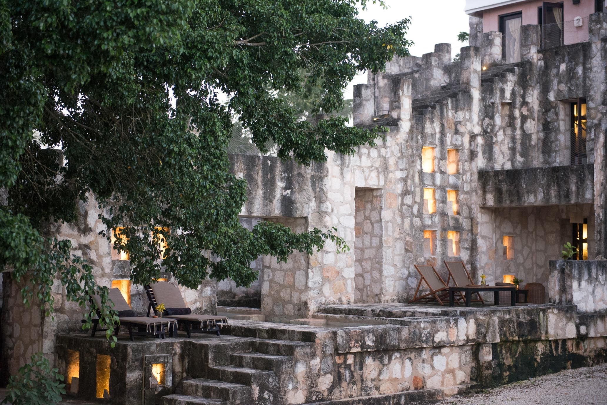 Coqui Coqui Papholchac, Mexico — Stone courtyard at dusk with ivy-draped walls, glowing windows, and wooden lounge chairs on terraced levels.