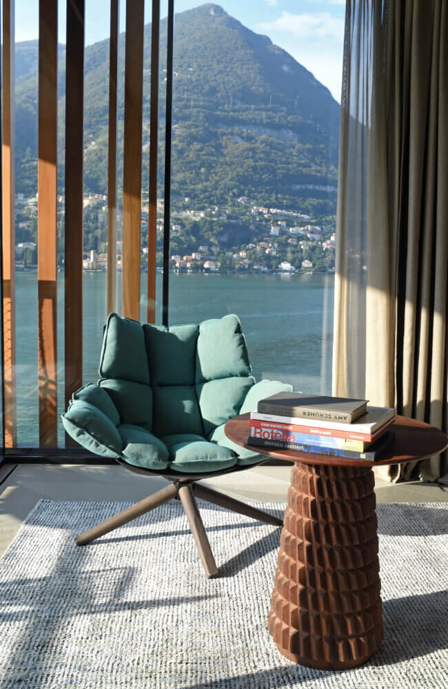 il Sereno Lago di Como, Italy — Minimalist bedroom chair with lake and alpine mountain views through floor-to-ceiling windows, mid-century modern side table with books, natural light, contemporary luxury interior design.