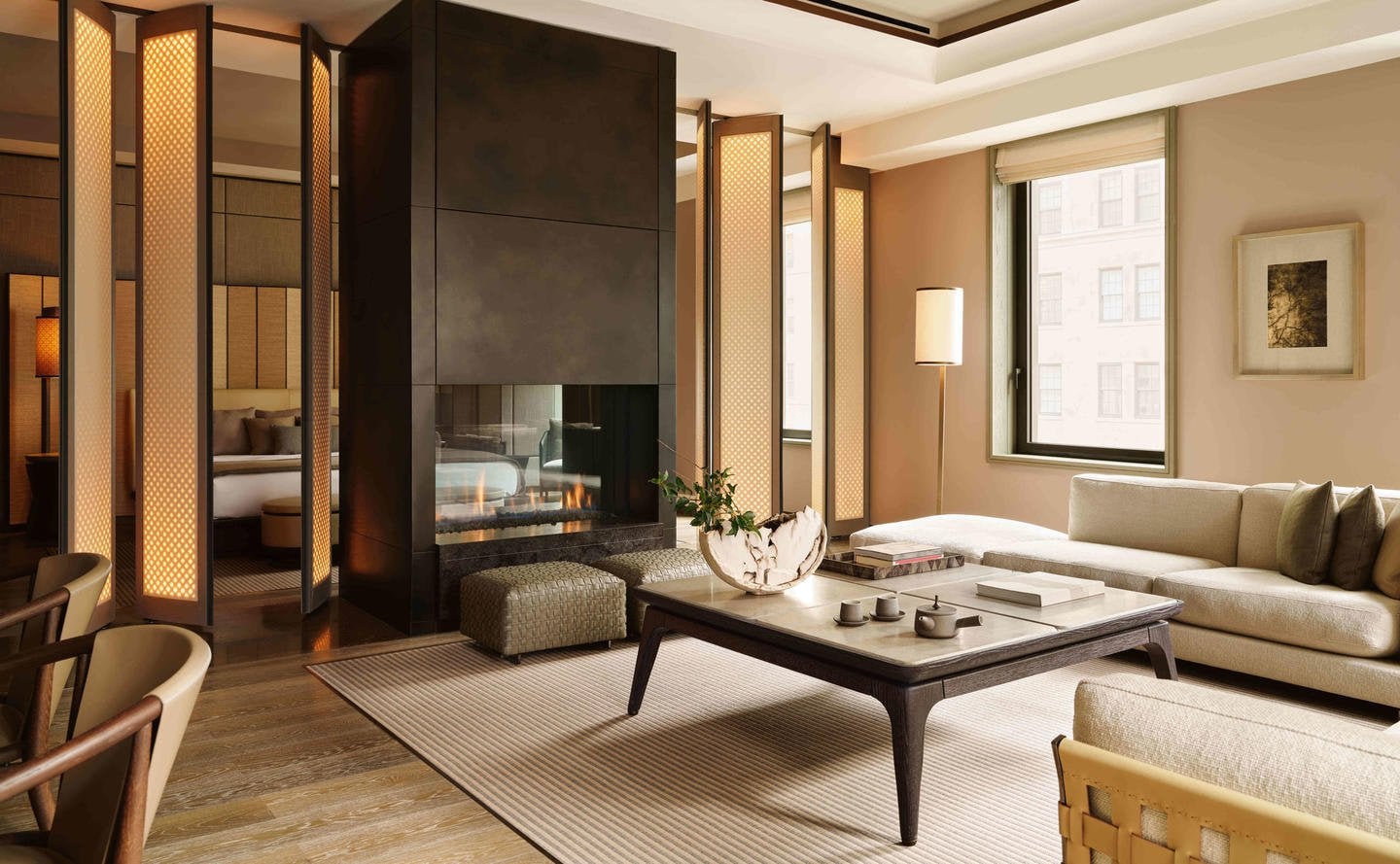 Aman New York, Manhattan, United States — Contemporary suite with fireplace, cream sectional sofa, dark wood screen dividers, and warm ambient lighting overlooking city.