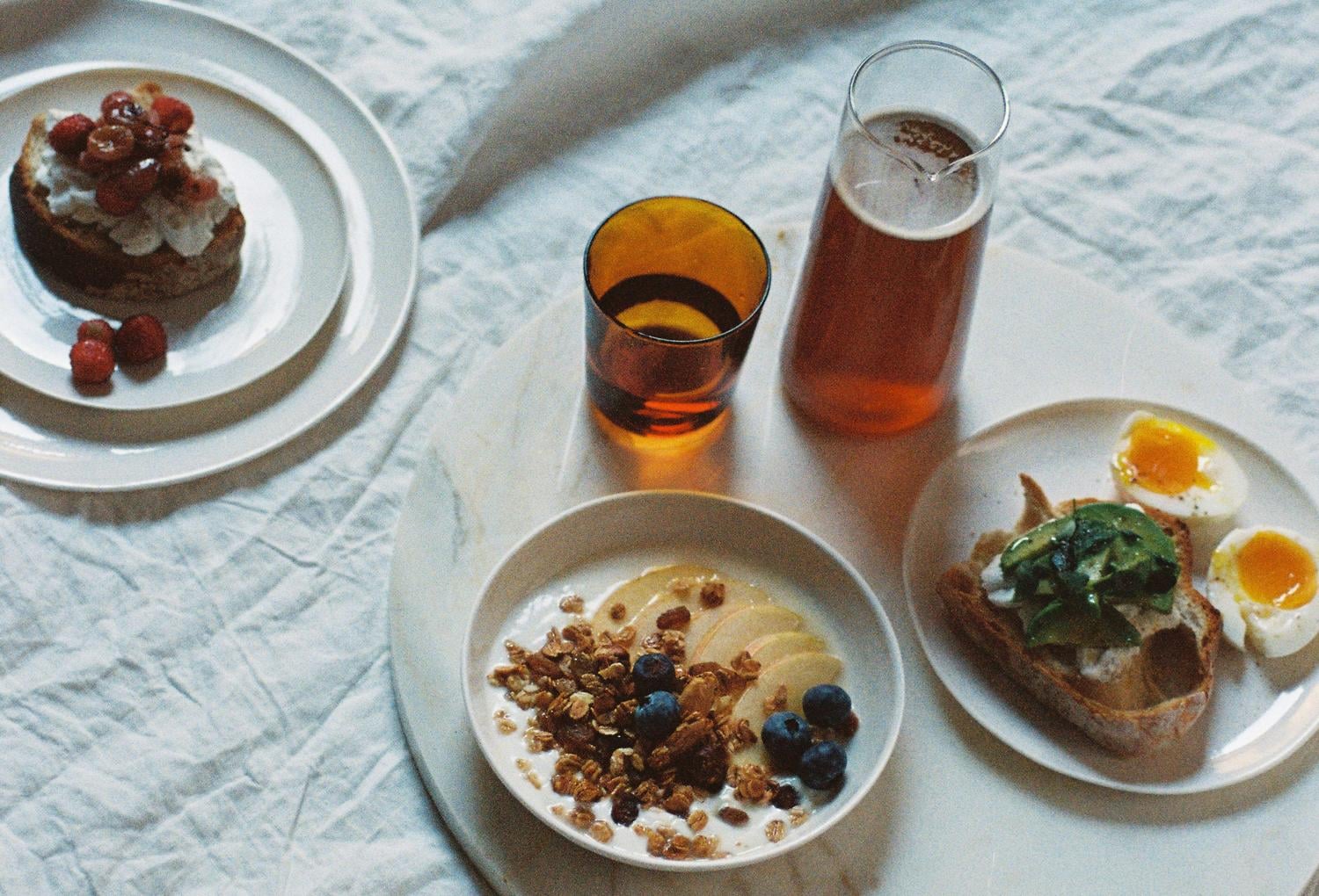 Courtyard Breakfast, Europe — Overhead shot of morning table with granola bowl, soft-boiled eggs on toast, chocolate pastry, amber beverages, and white linens in natural light.