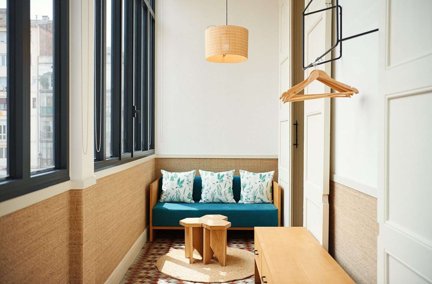 Casa Bonay, Europe — Minimalist hotel room with teal sofa, natural wood furniture, woven pendant light, and floor-to-ceiling windows overlooking city skyline.