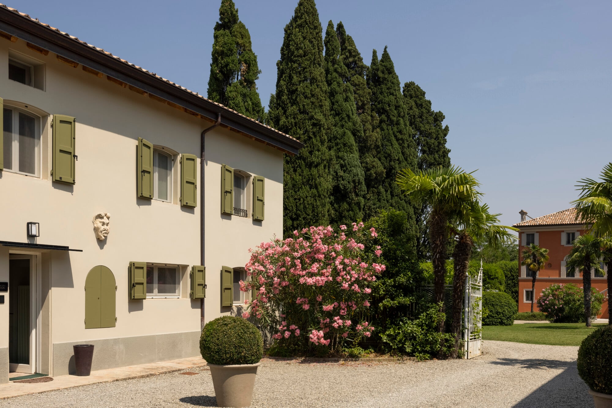Roncolo 1888, Europe — Cream-rendered villa with green shutters, pink oleander blooms, cypress trees, and gravel courtyard in Italian countryside estate setting.