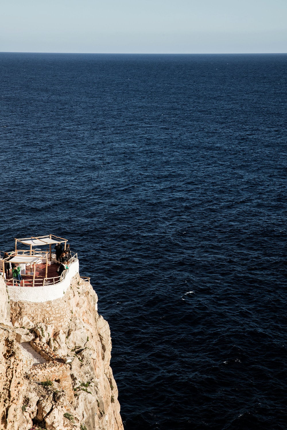 Cova d'en Xoroi, Balearic Islands — Whitewashed cliffside structure with wooden deck overlooking deep blue Mediterranean sea at horizon.