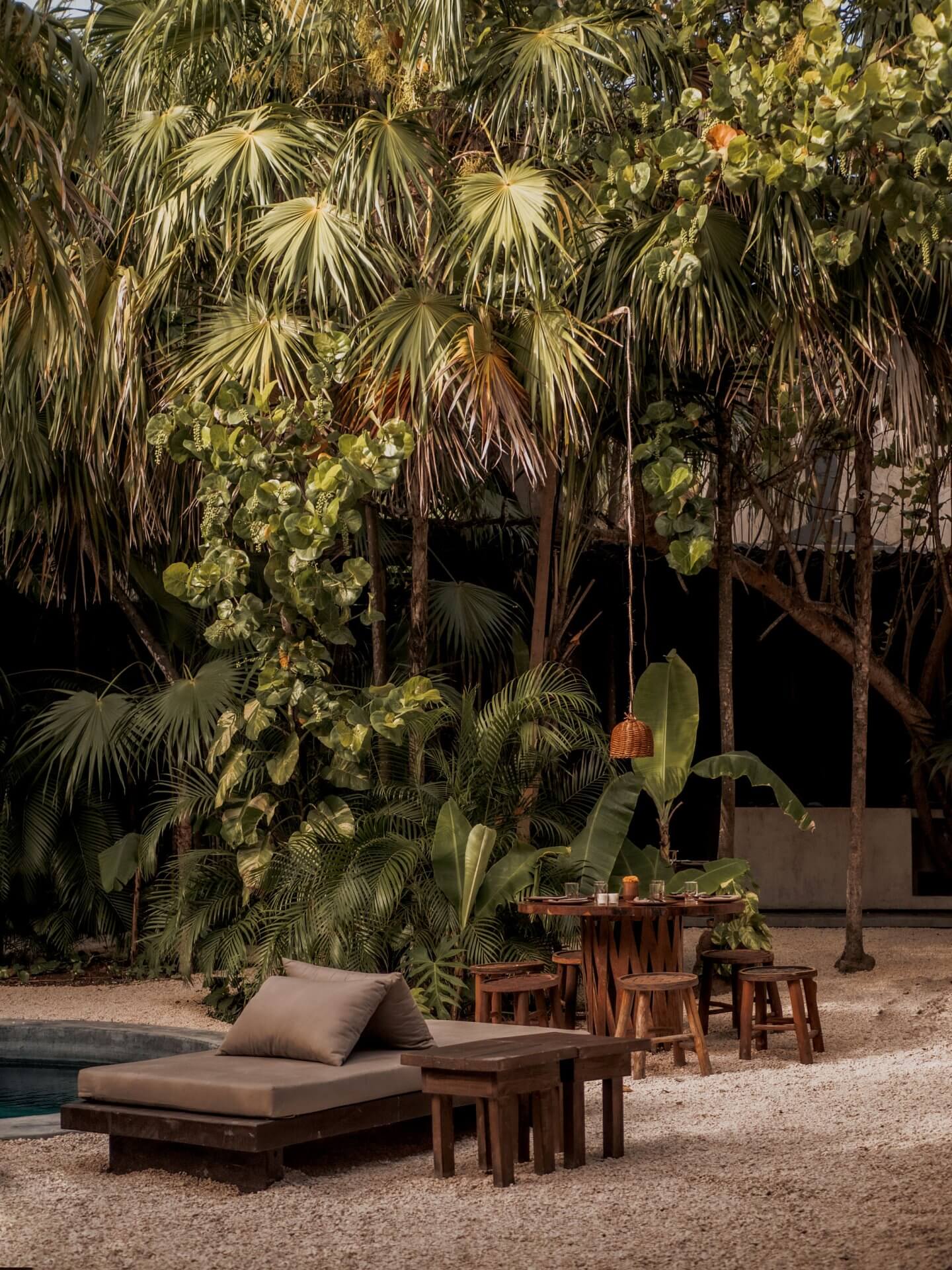 Tulum Treehouse, Mexico — Wooden lounge with cushions beneath towering palms and climbing vines, sandy courtyard at dusk with minimal furniture and tropical vegetation.