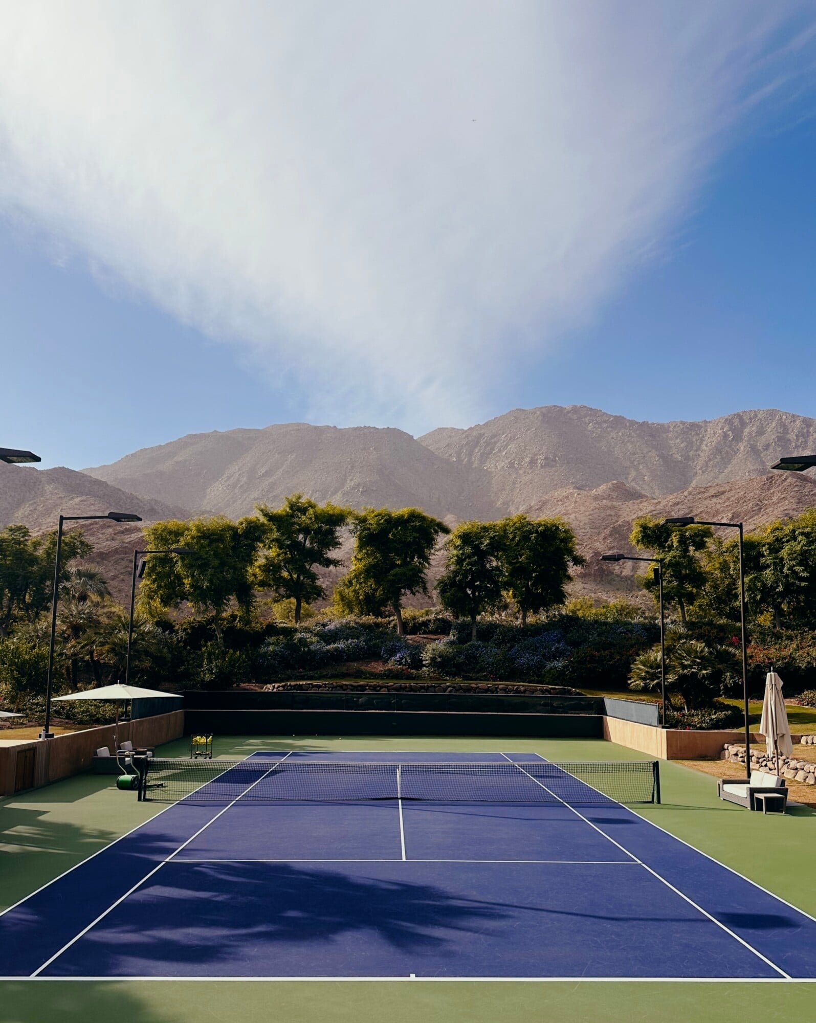 Tennis Lessons — Professional tennis court with blue and green hard courts, mountain views, tall trees, and modern lighting structures in arid landscape setting.
