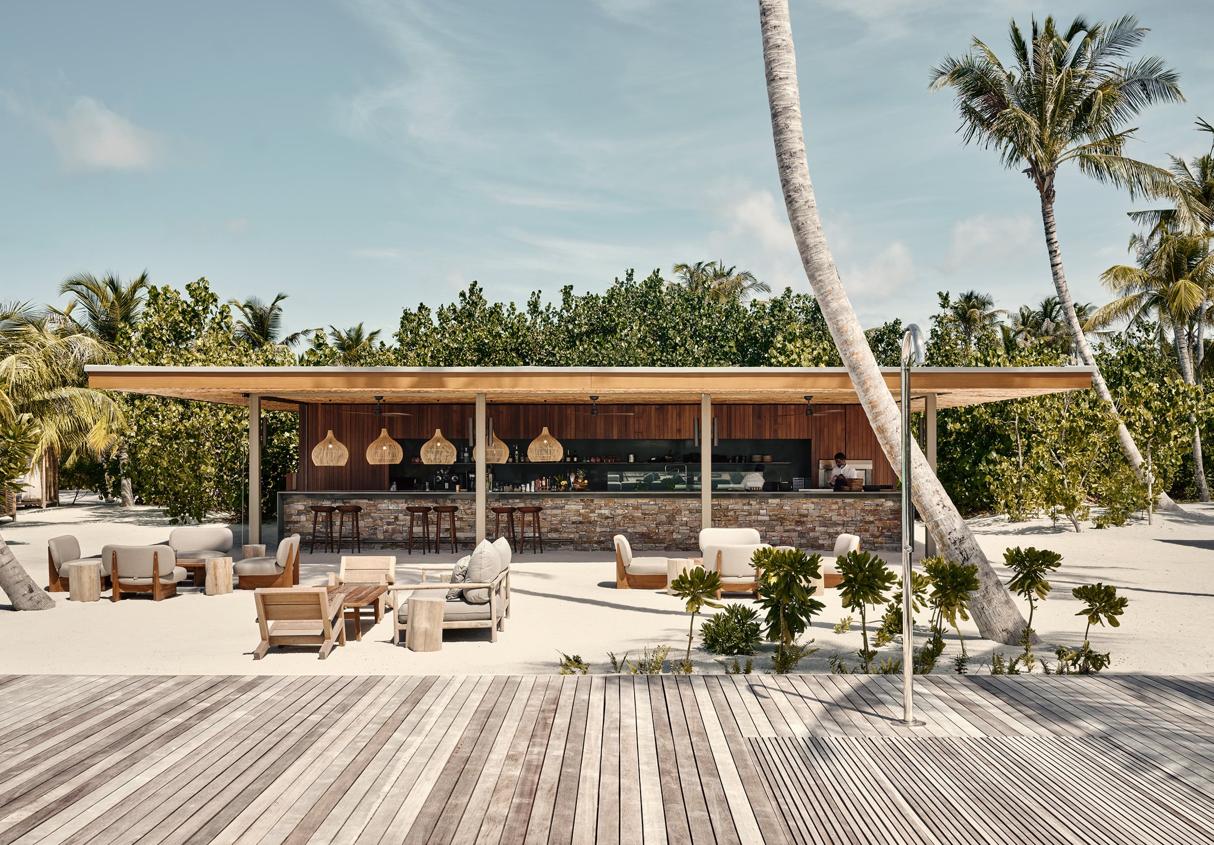 Patina Maldives, Asia — Beach bar with wooden pergola, stone counter, woven pendant lights, teak lounge chairs, palm trees, white sand deck under tropical sky.