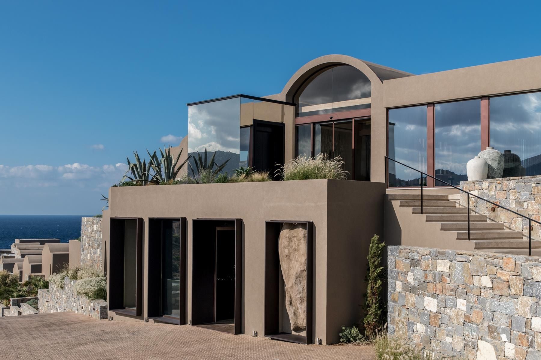 ACRO Suites, Europe — Modernist coastal architecture with curved stone facade, terracotta planter, black-framed glazing, natural materials, Mediterranean sea view.