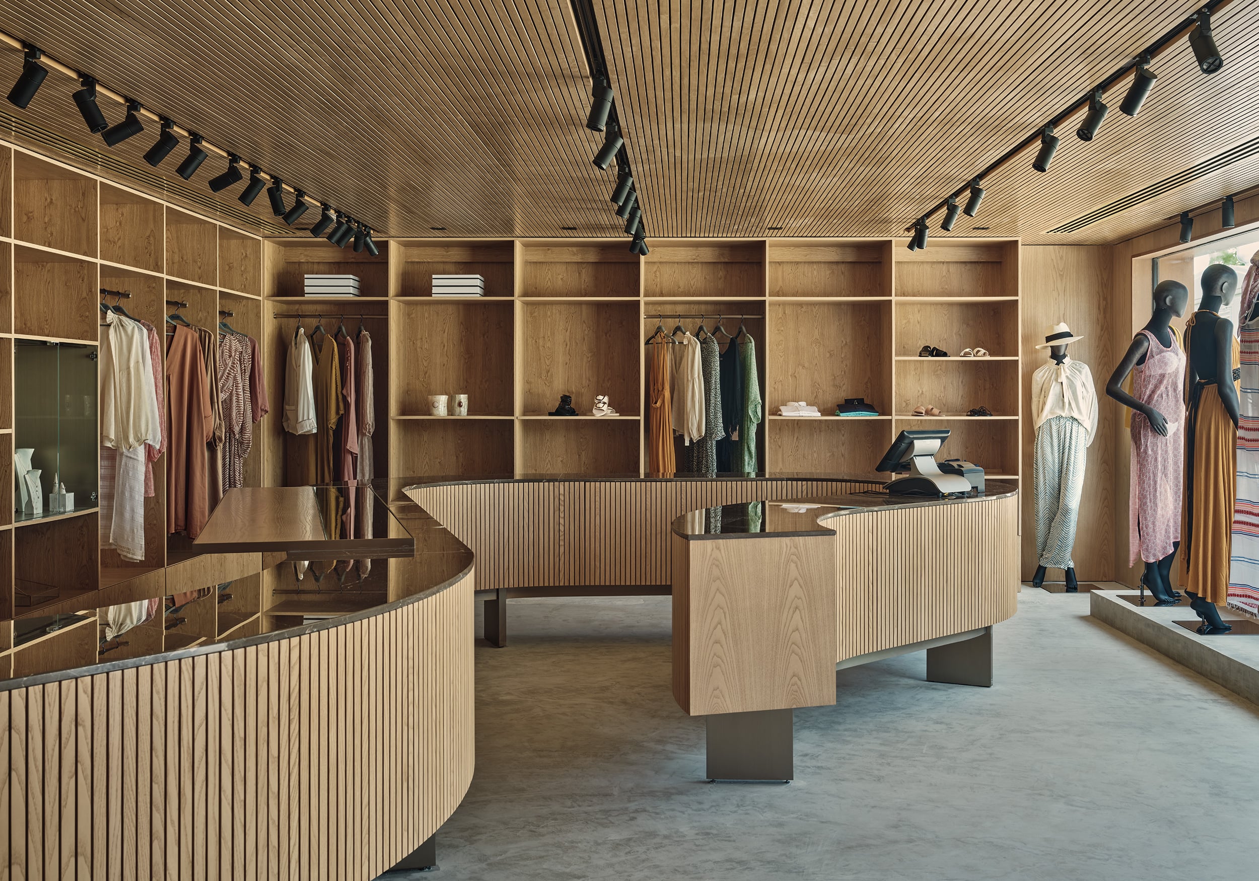 Patina Maldives, Asia — Wooden retail interior with curved display counters, floor-to-ceiling shelving, clothing racks, and mannequins showcasing luxury fashion collection under linear ceiling details.