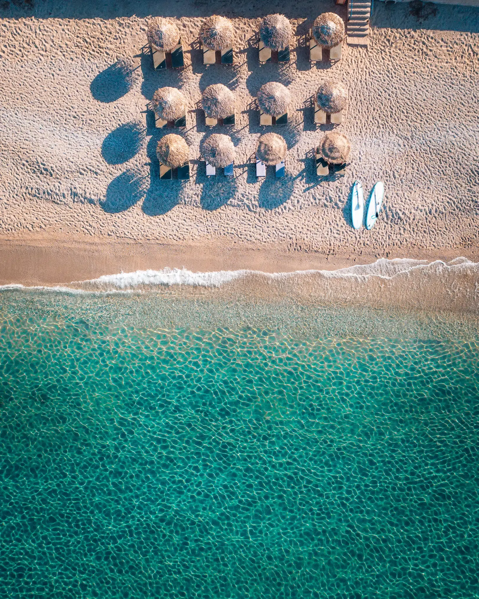 Water Sports & Lourdas Beach — Aerial view of organized beach umbrellas and loungers casting shadows on sand beside turquoise Mediterranean waters with white waves and surfboards.