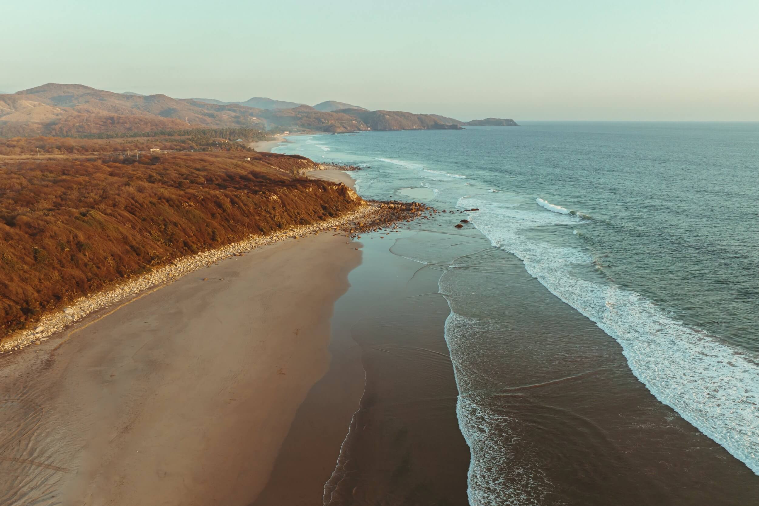 Hotelito MUSA, Mexico — Coastal cliffs overlook a pristine beach where turquoise waves meet golden sand, with rocky headlands extending into calm waters under hazy light.
