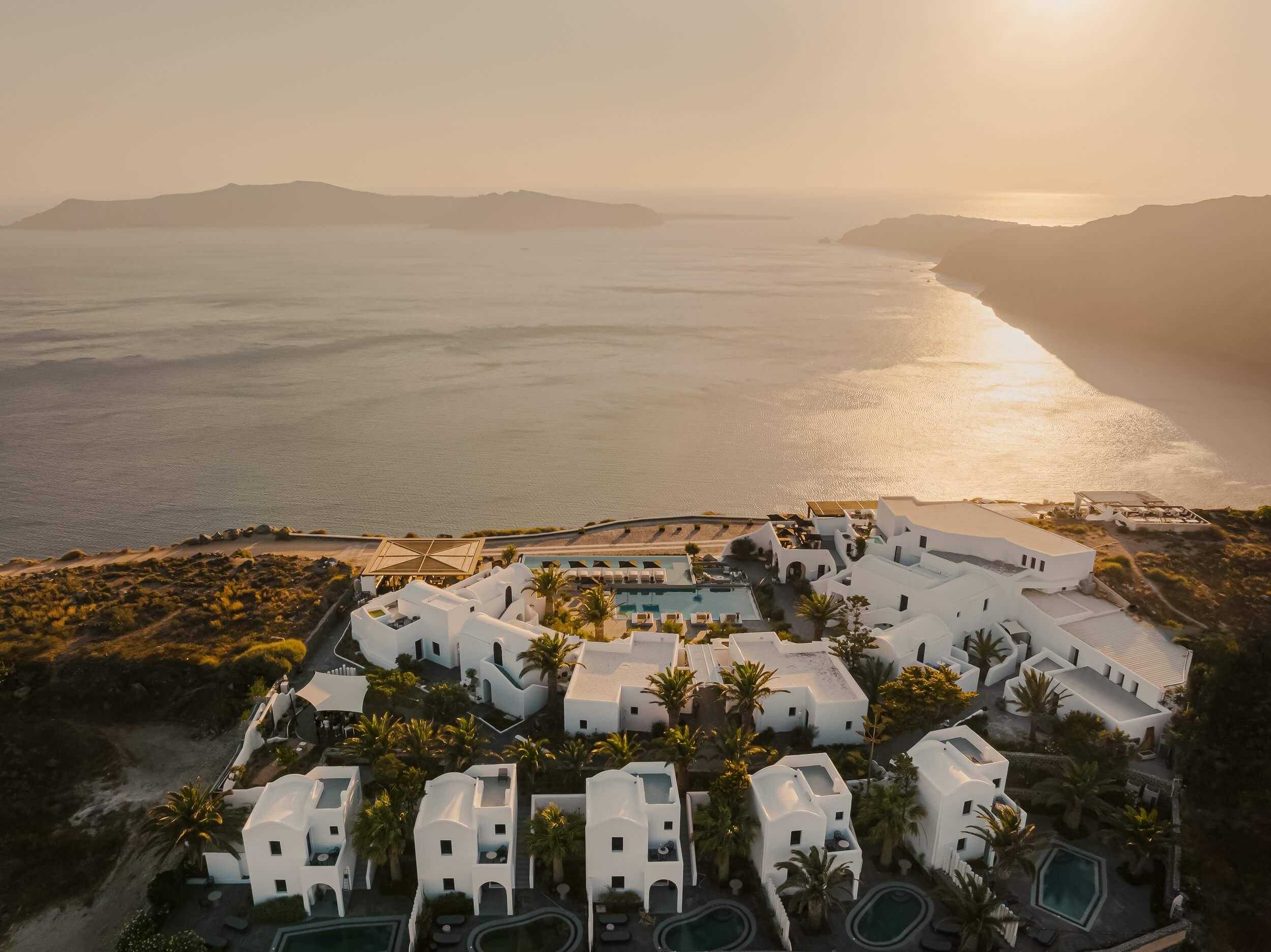 Nobu Santorini, Greece — Whitewashed Cycladic architecture with infinity pools overlooking caldera at golden hour, palm trees and volcanic islands in distant haze.