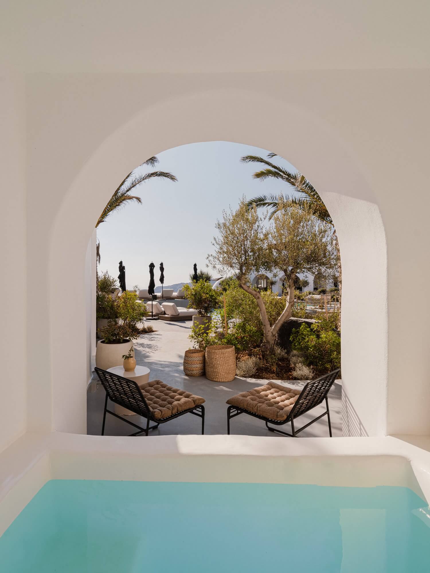 Nobu Santorini, Greece — Arched white aperture framing turquoise pool edge, olive trees, woven baskets, and terracotta chairs overlooking Aegean landscape.