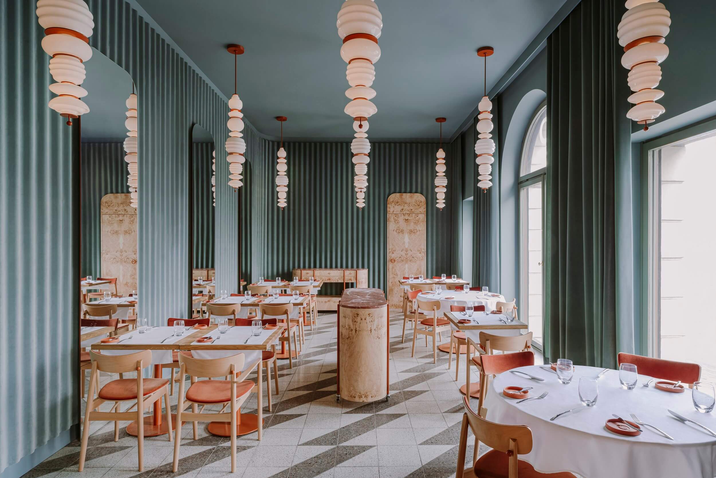 Opasly Tom, Europe — Modern restaurant interior with sage-green corrugated walls, stacked disc pendant lights, natural wood chairs, terrazzo flooring, and minimalist table settings.