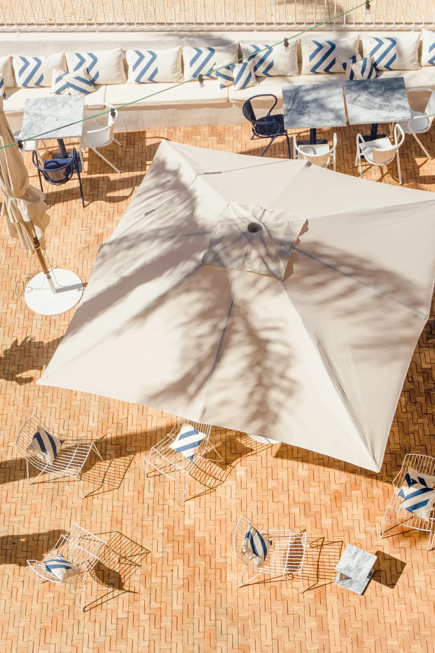 Casa Mãe, Europe — Overhead view of cream parasol with dappled shadow, woven loungers with blue striped cushions, terracotta tile courtyard, minimalist luxury hospitality design.