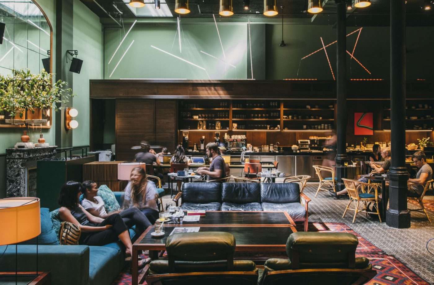 Libertine, Europe — Modern hospitality lounge with sage green walls, pendant lighting, black leather seating, wooden bar, neon signage, and guests working and socializing.