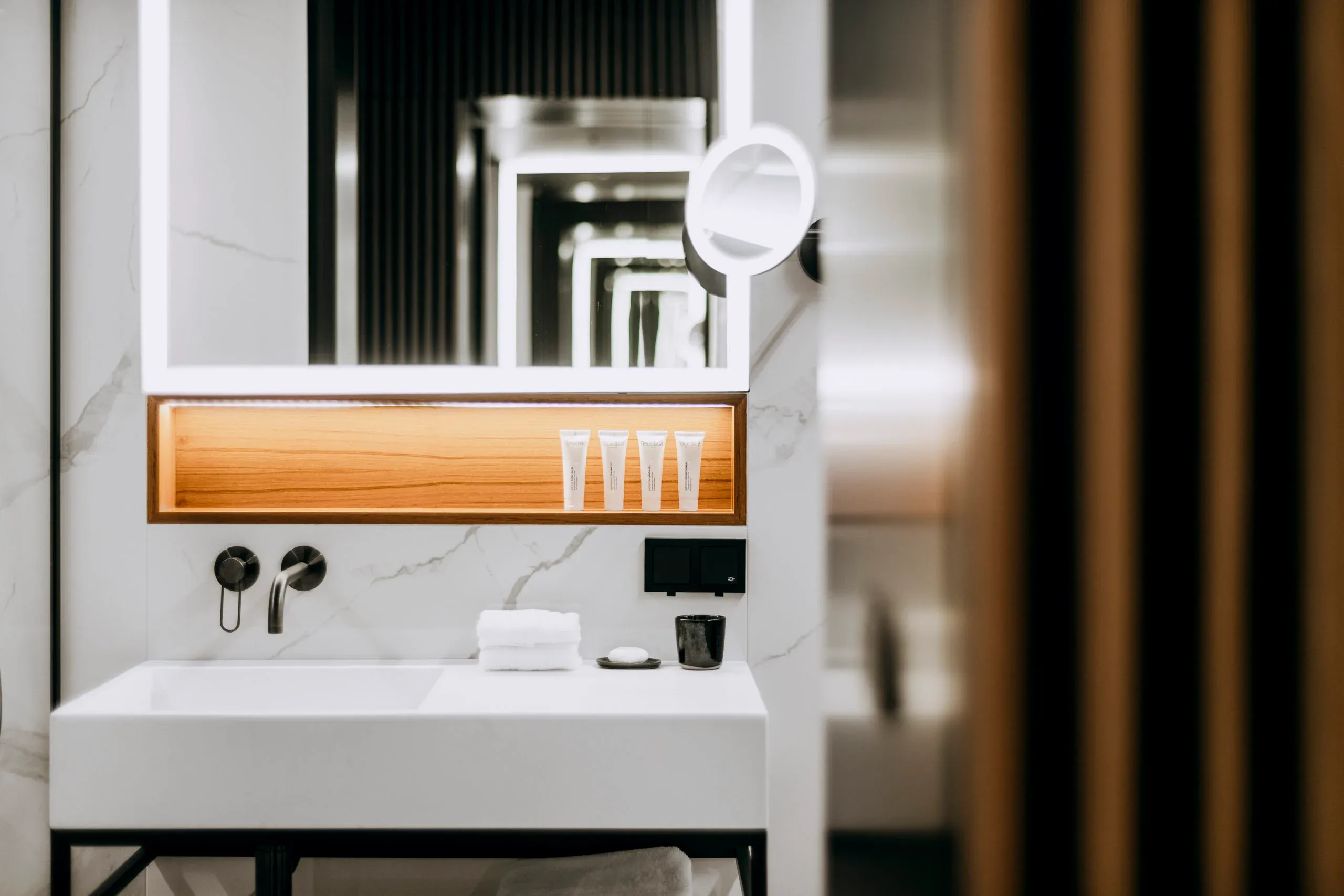 Nobu Warsaw, Poland — Modern bathroom with marble walls, wooden shelf, white vessel sink, and illuminated mirror reflecting contemporary luxury hotel design.