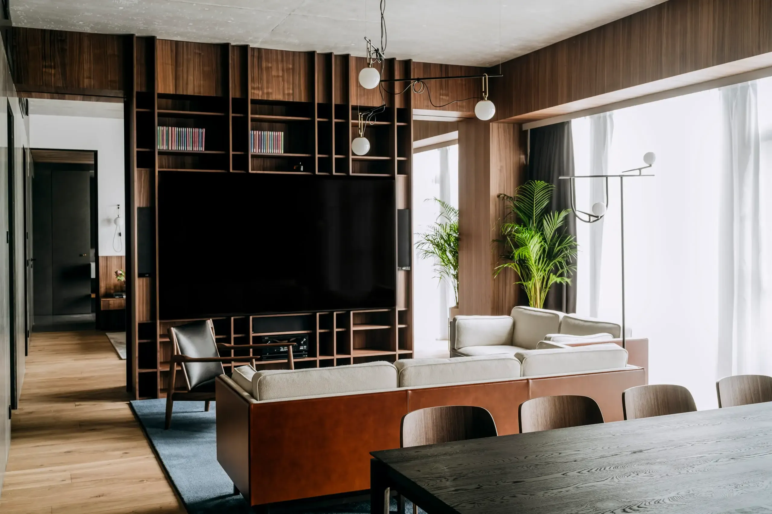 Nobu Warsaw, Poland — Modern living room with walnut wall paneling, linear shelving, large flat-screen TV, cream sectional sofa, pendant lights, and floor-to-ceiling windows with city views.