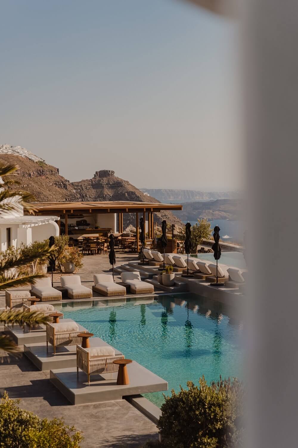 Nobu Santorini, Greece — Clifftop pool with turquoise water, wooden pavilion, loungers, cypress trees, caldera views at golden hour.