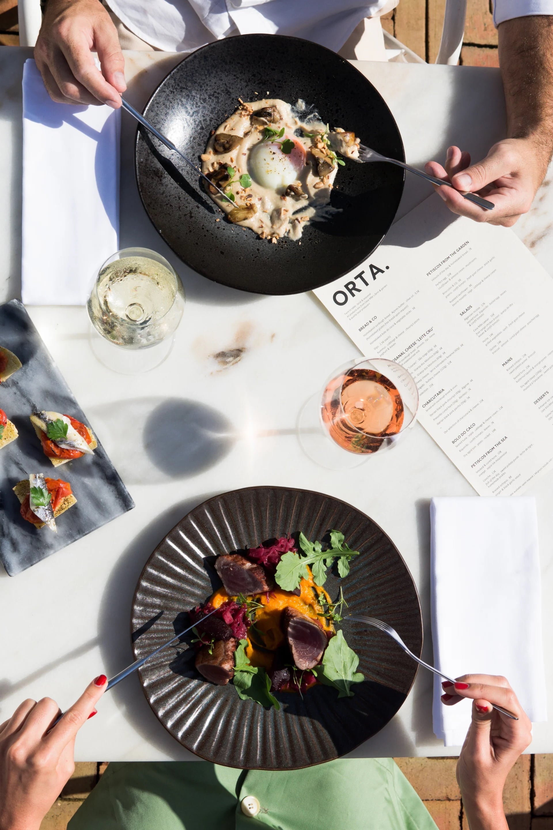 Casa Mãe, Europe — Overhead dining scene with plated dishes, wine glasses, and menu on white table in sunlight, contemporary fine dining.