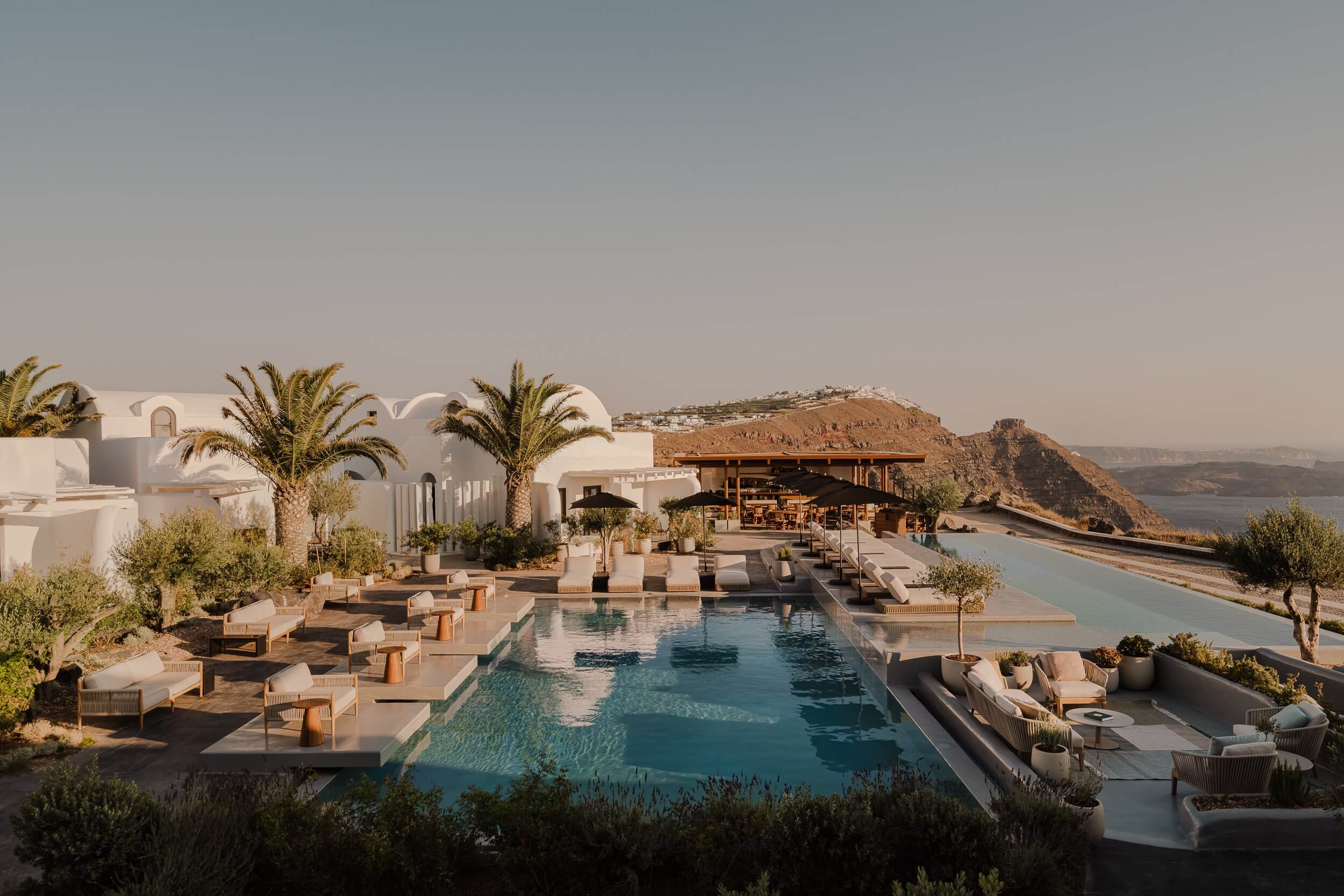 Nobu Santorini, Santorini, Greece — whitewashed architecture with infinity pool overlooking caldera, teak loungers, palm trees, and volcanic landscape at golden hour.