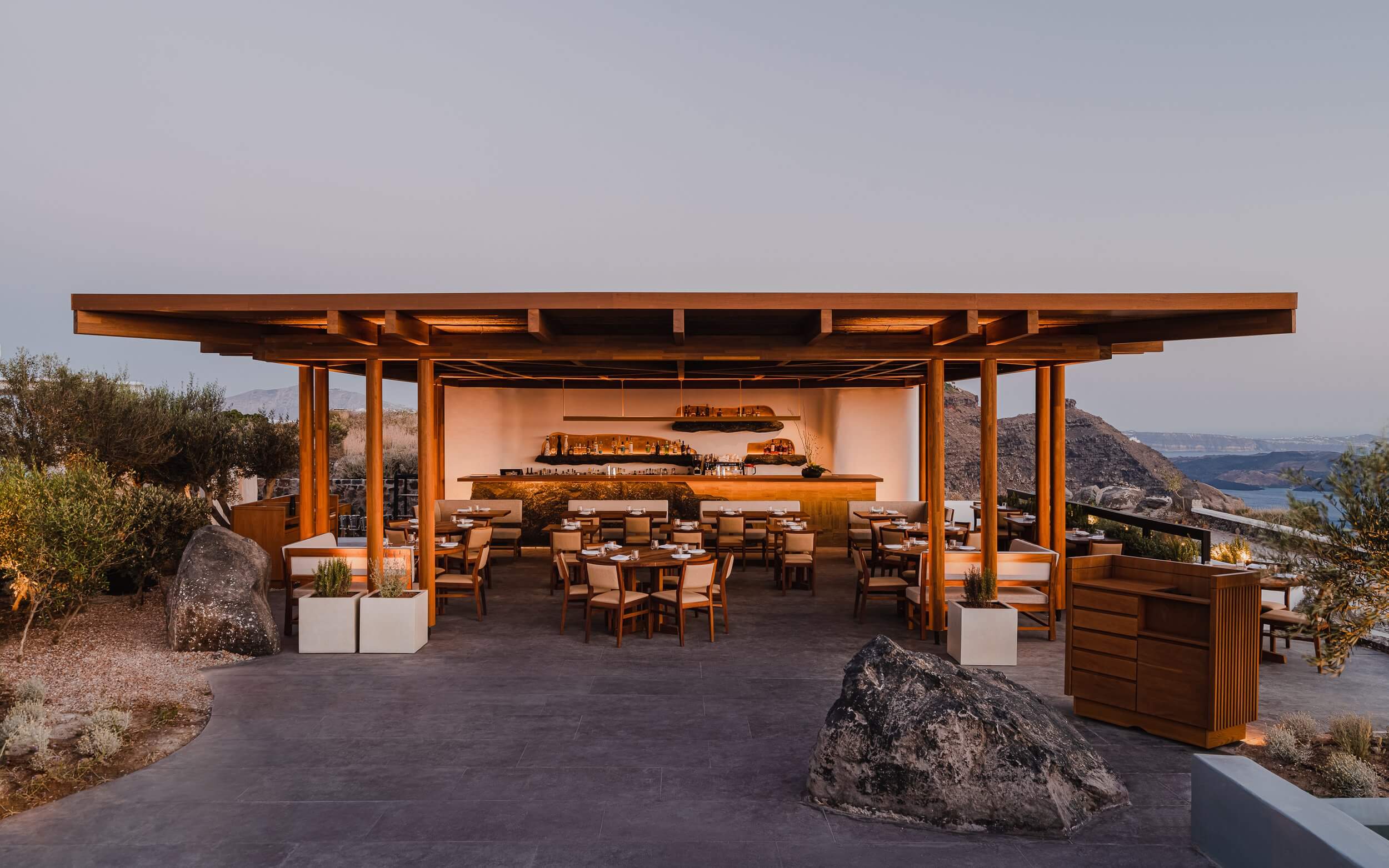 Nobu Santorini, Europe — Open-air restaurant pavilion with timber pergola, bar counter, Cycladic sea views, natural stone elements, warm ambient lighting at dusk.