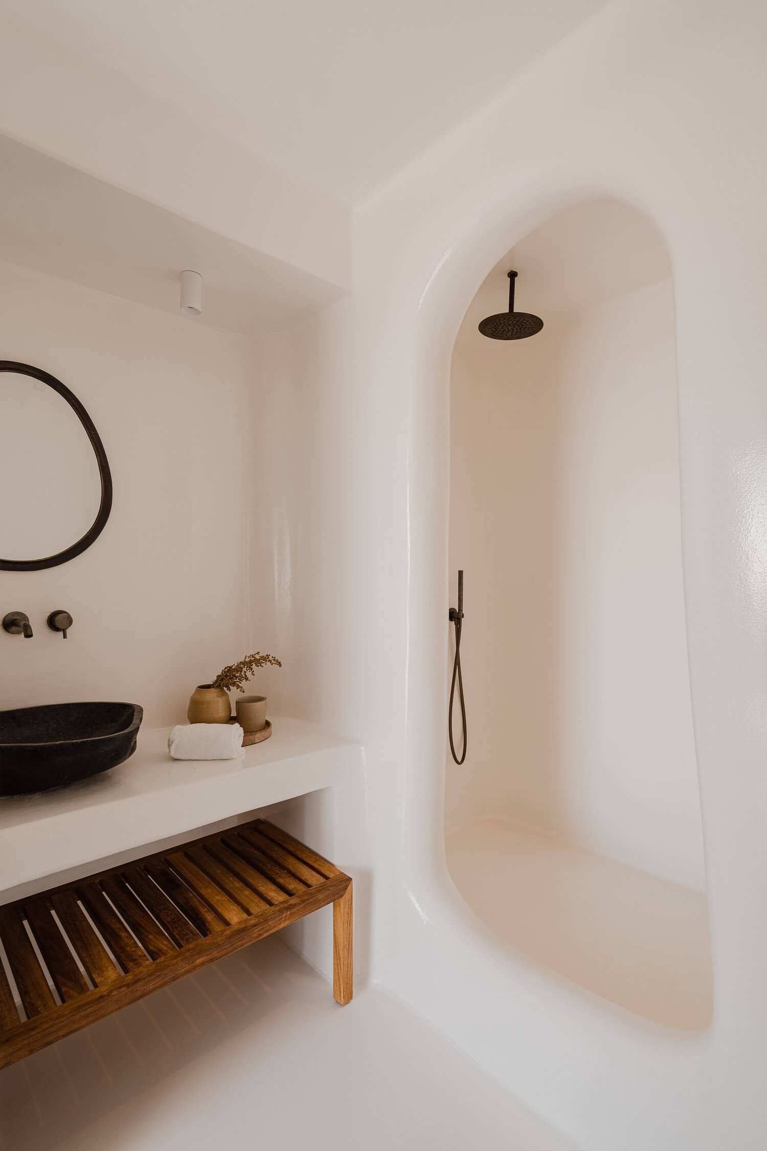 Nobu Santorini, Europe — Minimalist bathroom with arched alcove shower, black fixtures, curved white walls, wooden bench, and black vessel sink.