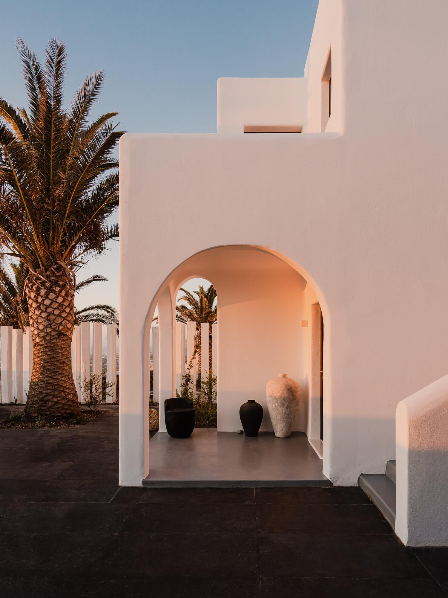 Nobu Santorini, Europe — Minimalist white archway with curved aperture, terracotta vessels, palm tree, golden hour light, Mediterranean luxury design.