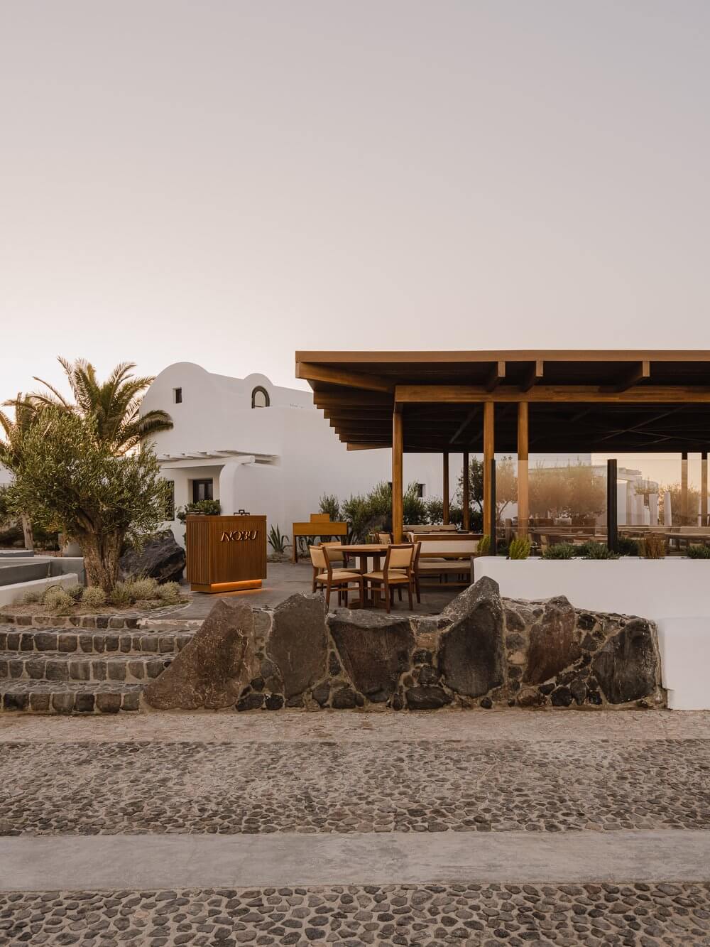 Nobu Santorini, Greece — Whitewashed Cycladic architecture with wooden pergola dining pavilion overlooking beach, natural stone seating, and palm landscaping at sunset.