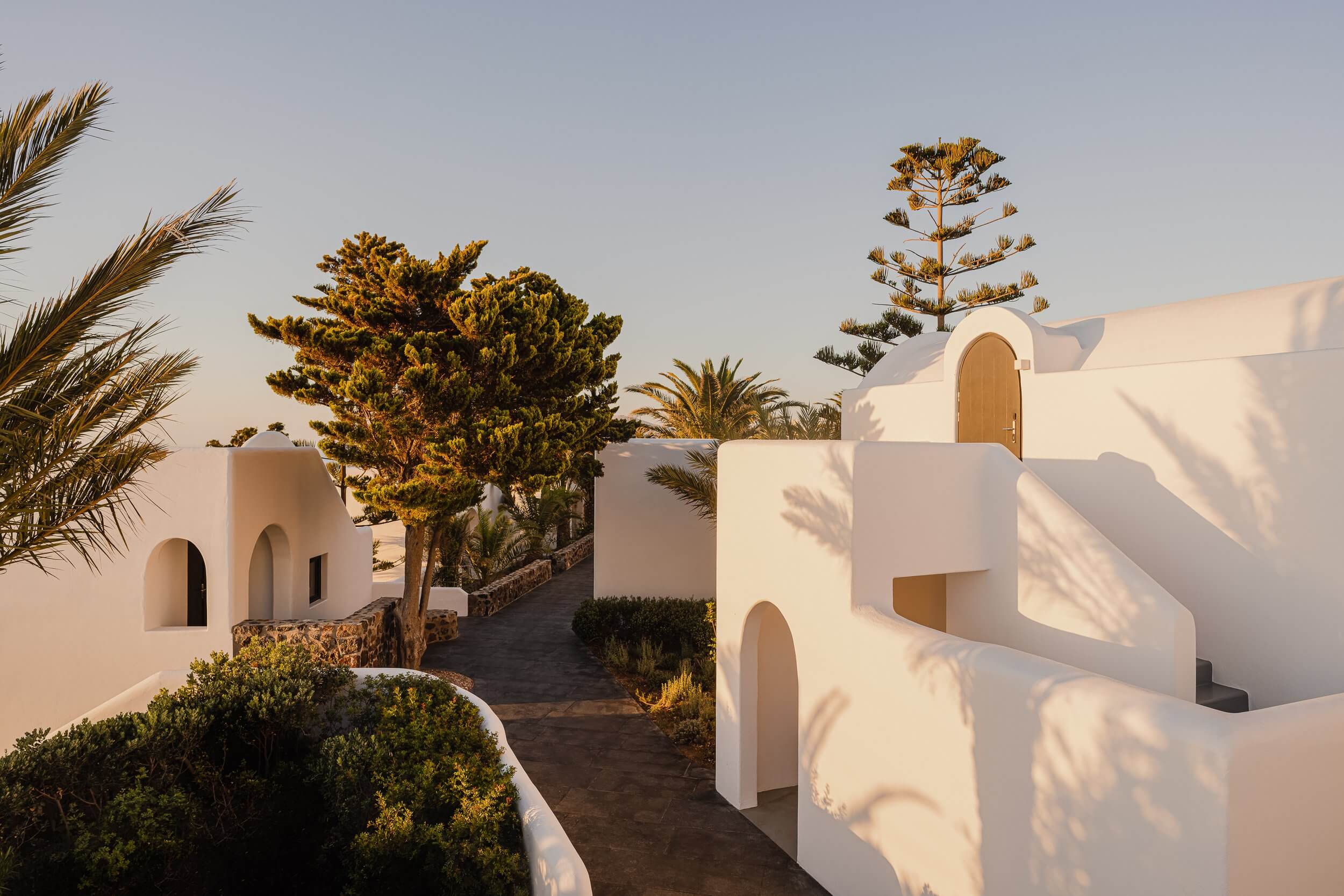 Nobu Santorini, Europe — Whitewashed Cycladic architecture with curved walls, arched doorways, and palm trees casting shadows in golden sunlight.