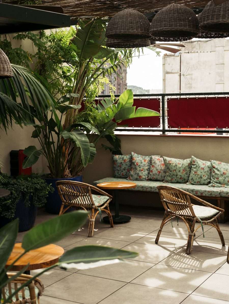 Casa Bonay, Europe — Rooftop terrace with woven pendant lights, rattan chairs, flowering cushioned bench, tropical plants, and city views through maroon railings.