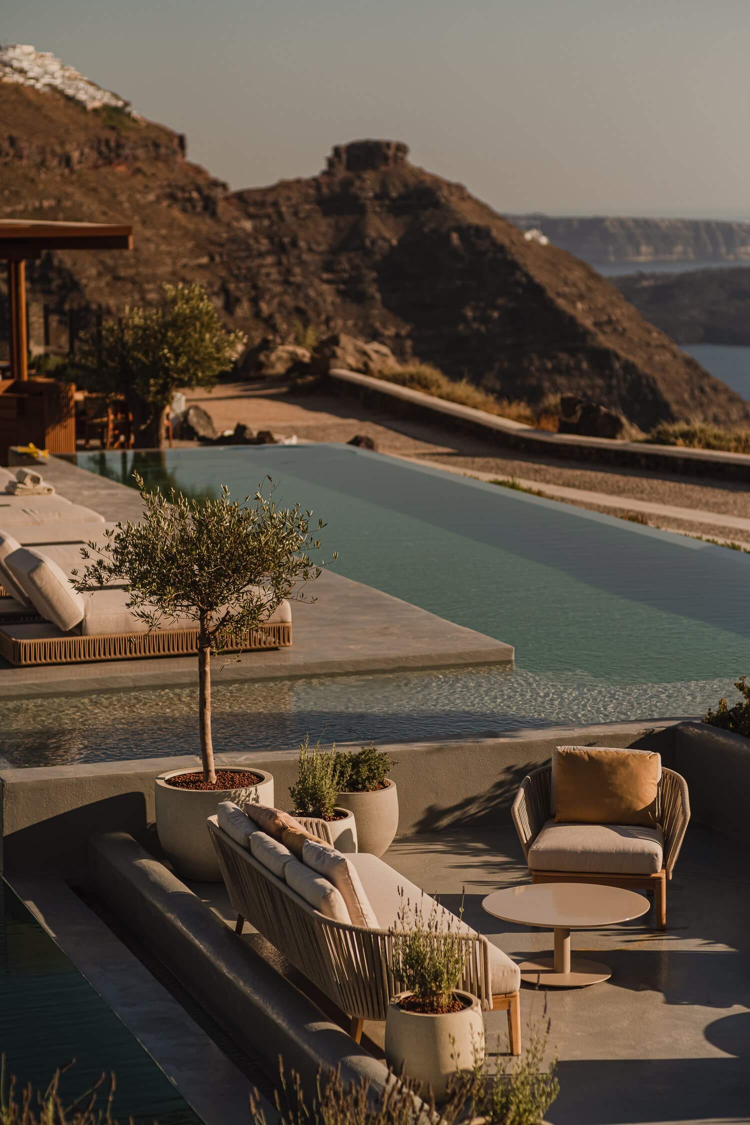 Nobu Santorini, Greece — Clifftop terrace with infinity pool, travertine loungers, olive trees, and caldera views at sunset.