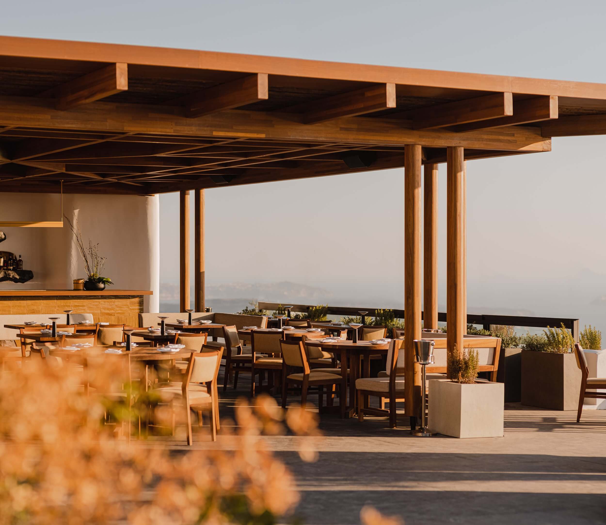 Nobu Santorini, Greece — Wooden pergola restaurant terrace with natural light, minimalist furnishings, caldera views, contemporary hospitality design.