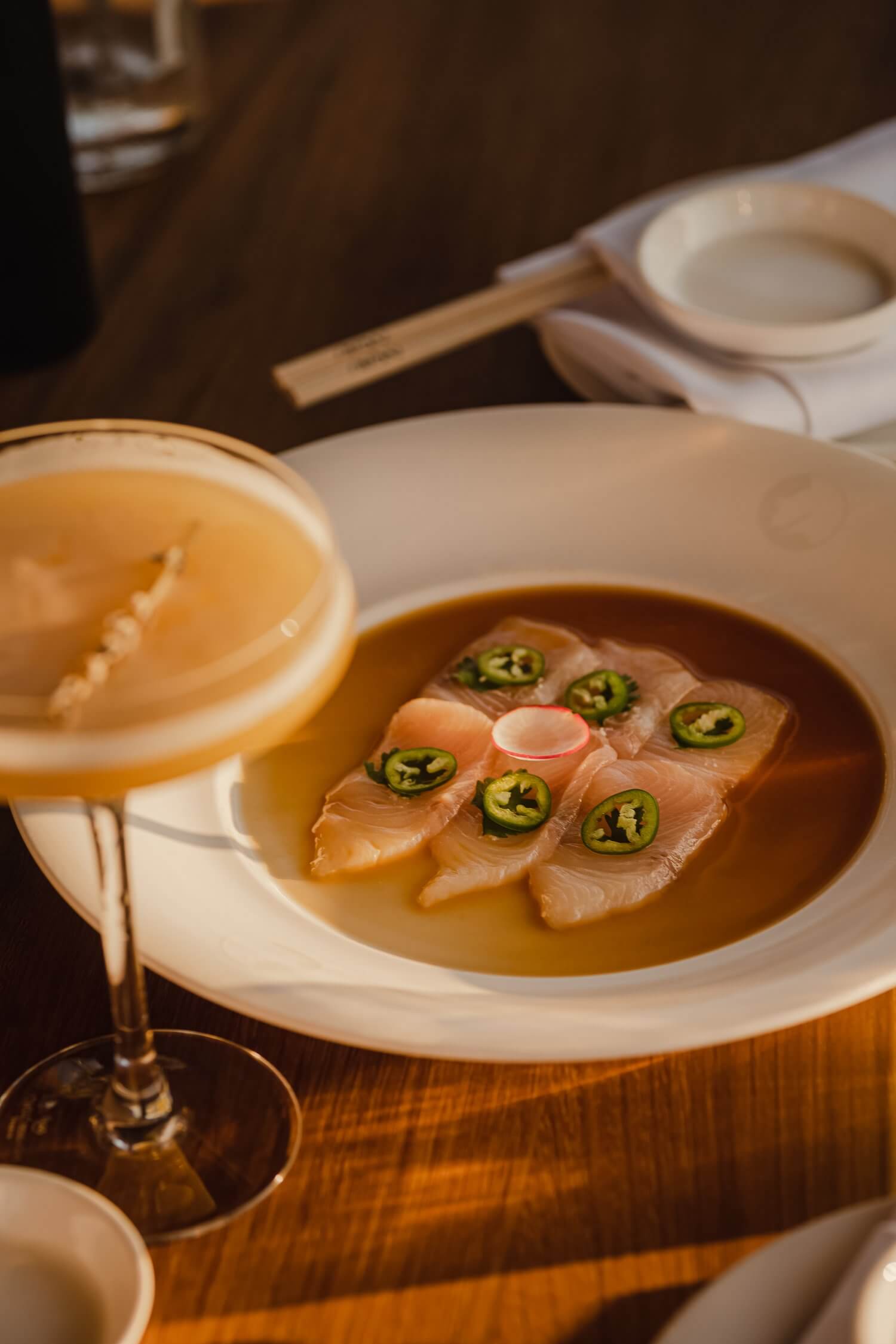 Nobu Restaurant, Europe — Seared fish with jalapeño garnish in dashi broth, served with sake in warm minimalist dining setting.