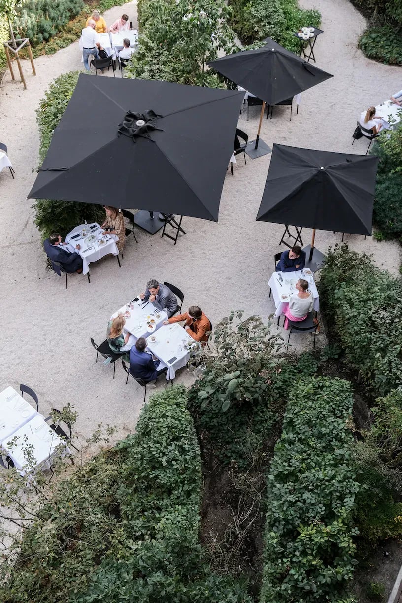 August, Europe — Overhead view of outdoor courtyard with black umbrellas sheltering diners at white-clothed tables surrounded by hedged landscaping and gravel pathways.