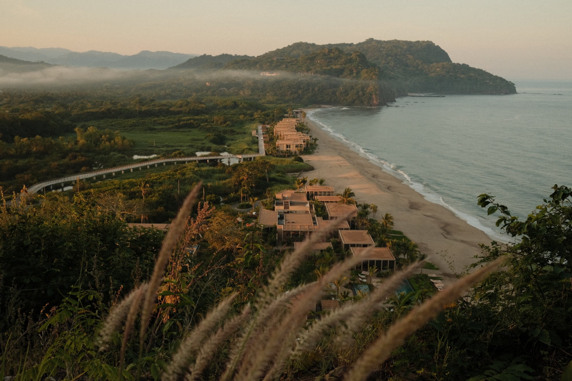 Rosewood Mandarina, Mexico — Clifftop resort villas with wooden architecture overlooking pristine beach, forested headlands, and morning mist in golden light.