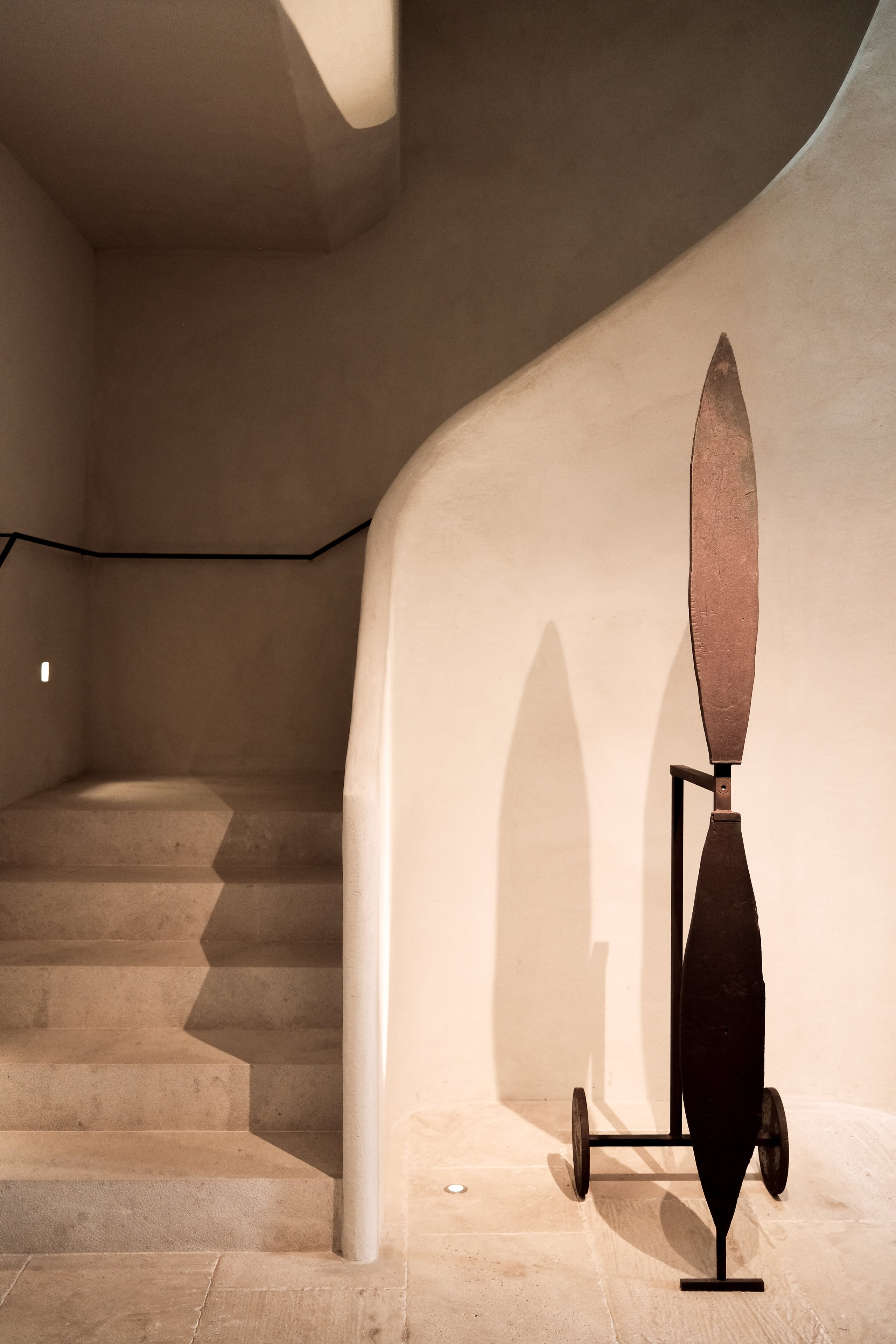 Can Ferrereta, Balearic Islands, Spain — Minimalist gallery space with curved white walls, stone stairs, sculptural bronze knife installation, warm natural light casting shadows.